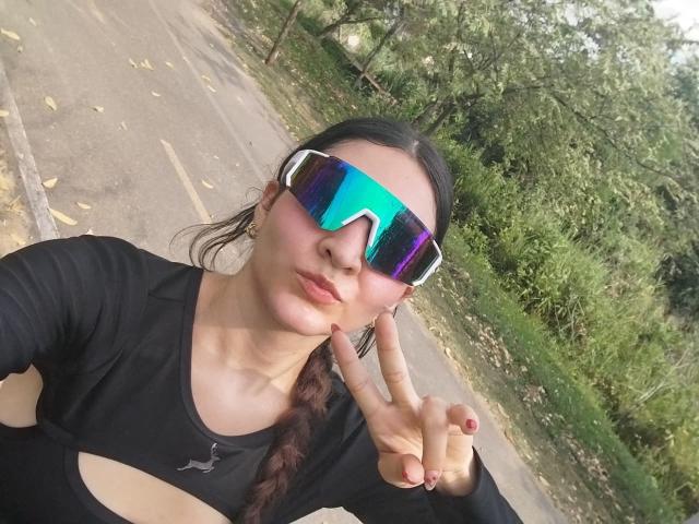 SeleneRoussel with dark hair in a braid, wearing reflective blue-green sunglasses, black sports top, and making a peace sign, showing her nose and lips.
