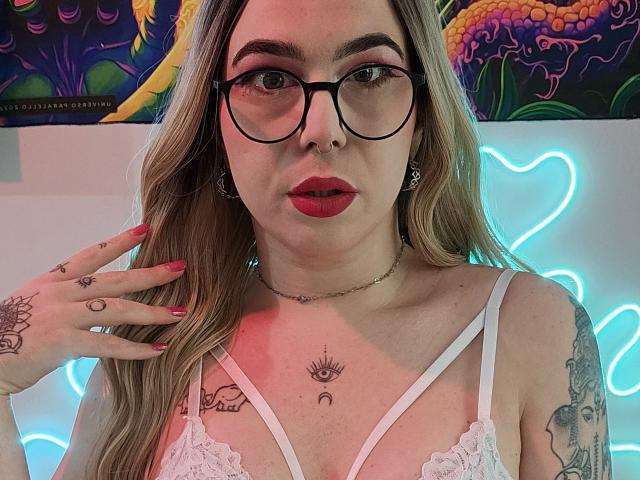 VickSexy with glasses, red lipstick, white lace bra, tattoos on chest and hand, long blonde hair, hand on neck, neon lights, and colorful wall art in background.