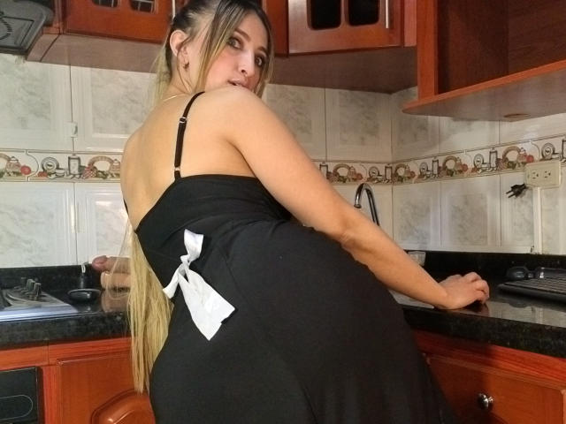 AnastasiaVolkov with long blonde hair, black dress with white bow, looking back, showing her bare back and black bra straps, standing in a kitchen with wooden cabinets.