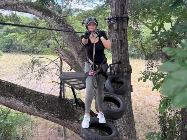 KatherinLatin, wearing a black shirt, gray pants, and white shoes, hangs from a tree by a rope, with tires and a metal bench in the background.