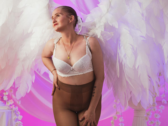 RubiAnderson, con un reggiseno di pizzo bianco, calze marroni e ali d'angelo bianche, guarda verso l'alto, indossa una collana d'oro e ha un tatuaggio sul braccio destro, in un ambiente illuminato di rosa..