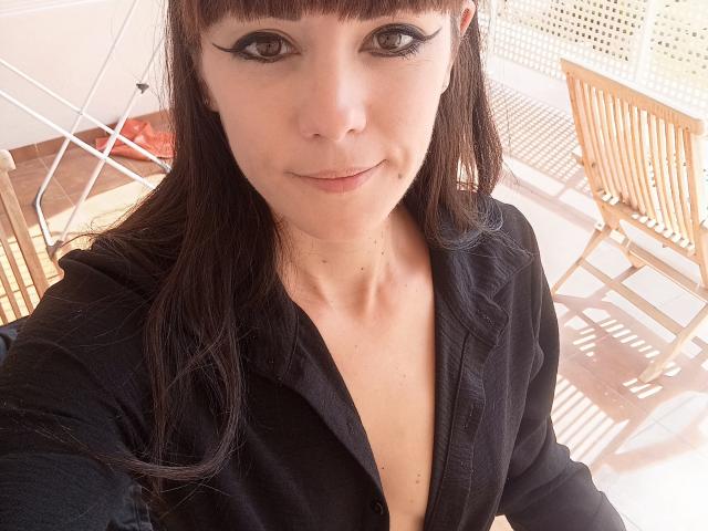 OrianaLaFrancaise with long brown hair, black eyeliner, black top, and a slight smile, showing a cute face and small tits, standing in a sunlit room.