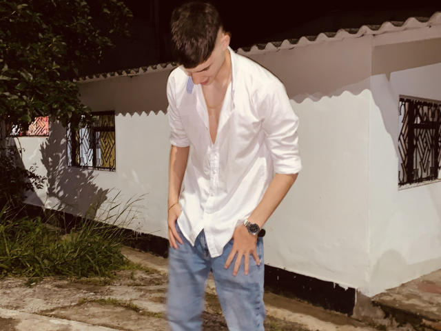 DrakeJonees, wearing a loose white shirt and blue jeans, bends over with hands on his butt, looking down. He has short dark hair and a black watch.