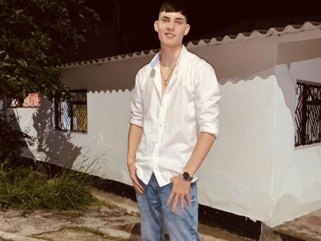 DrakeJonees stands with hands in jeans pockets, wearing a white button-up shirt, black watch, and necklace, smiling with short black hair, in a daytime outdoor setting.