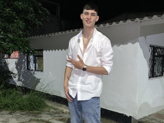 DrakeJonees, standing, wearing a white button-up shirt, blue jeans, and a watch, hand on chest, smiling, short brown hair, white skin, night, house with black barred windows.