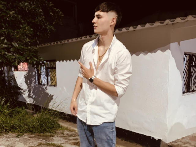 DrakeJonees, a slim young dude with a shaved side, wearing a white button-up shirt and blue jeans, stands outside with a hand on his chest, looking to the side.