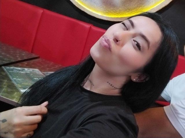 AlexaRosex with black hair, closed eyes, and pouty lips, wearing a black shirt, touching her shirt with one hand, in a red booth. Her ass is slightly visible.