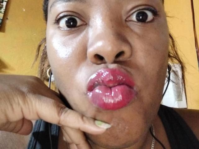 BrownieBabe with shiny brown skin, wide eyes, and glossy pink lips, making a pouty face. She's touching her lower lip with a green-nailled finger.