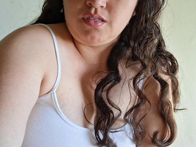 AlyceDina's upper body, showing her chubby face, wavy brown hair, and white tank top, with her small, perky tits slightly visible. She's wearing small earrings.