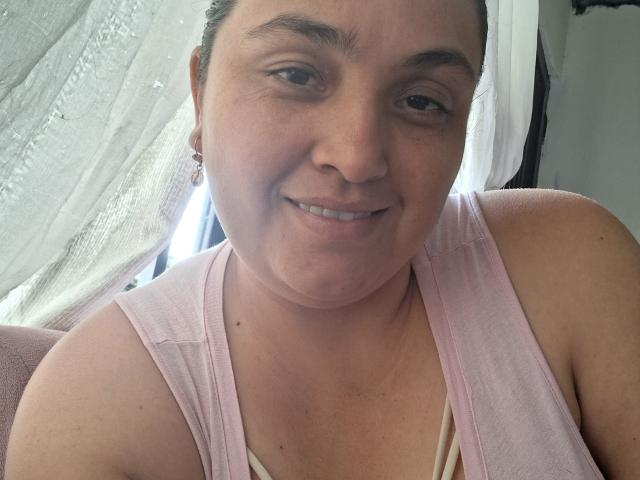 AlyceDina with short brown hair, smiling, wearing a pink tank top, showing her chubby face and small tits. She has a light skin tone and is indoors.