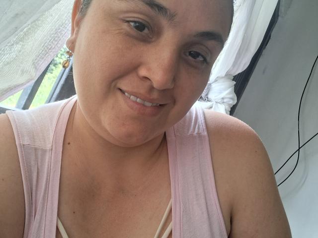 AlyceDina with light brown skin, small nose, and dark eyebrows, smiling softly. She's wearing a pink, sleeveless top, showing a slight cleavage. Her hair is tied back.
