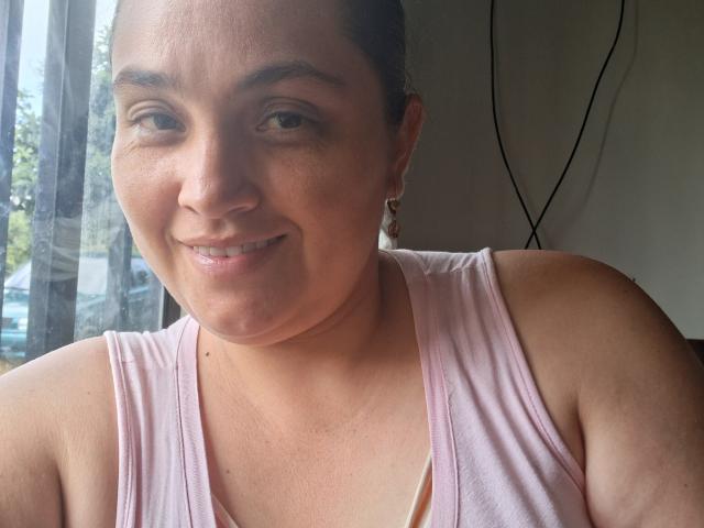 AlyceDina with medium brown skin, dark brown eyes, and a slight smile. She's wearing a pink tank top, showing her small tits and bare shoulders. Background is blurred.
