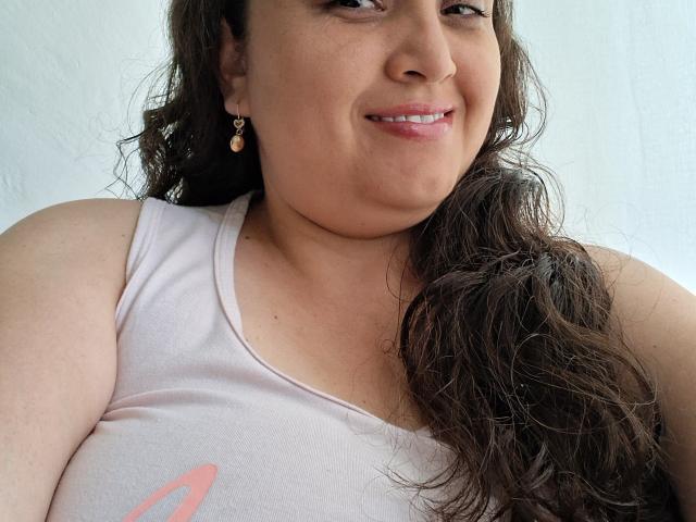 AlyceDina with wavy brown hair, light pink tank top, small pink earrings, smiling, showing her tits, light skin, pink lips, and a cute, relaxed look.
