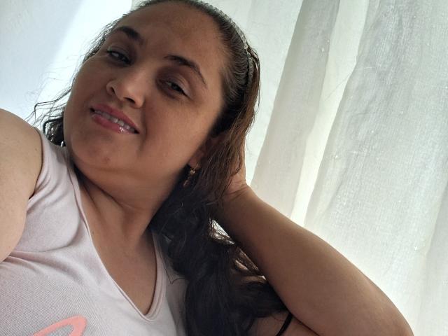 AlyceDina with wet brown hair, wearing a white tank top, smiling slightly, hand in hair, showing small pink nipple, light brown skin, casual selfie.