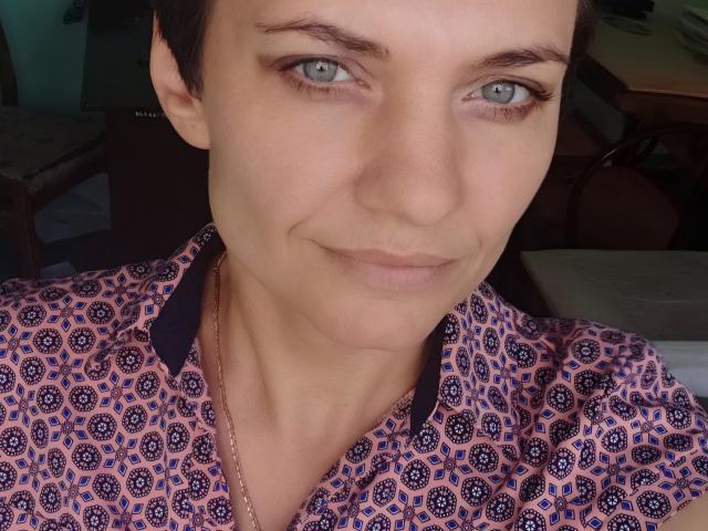 AvaSaturnian with short brown hair, blue eyes, and a slight smile, wearing a pink patterned shirt with black collars, showing her clean, pale skin and subtle makeup.