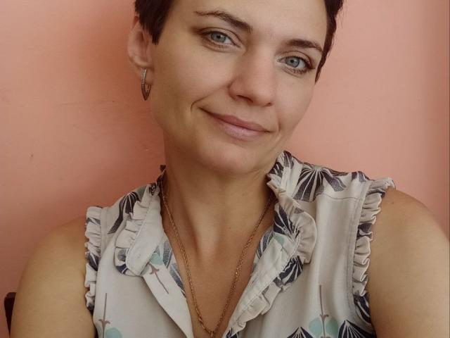 AvaSaturnian with short brown hair, small nose, and slight smile, wearing a floral sleeveless top, gold necklace, and hoop earrings, has blue eyes and a cute face.