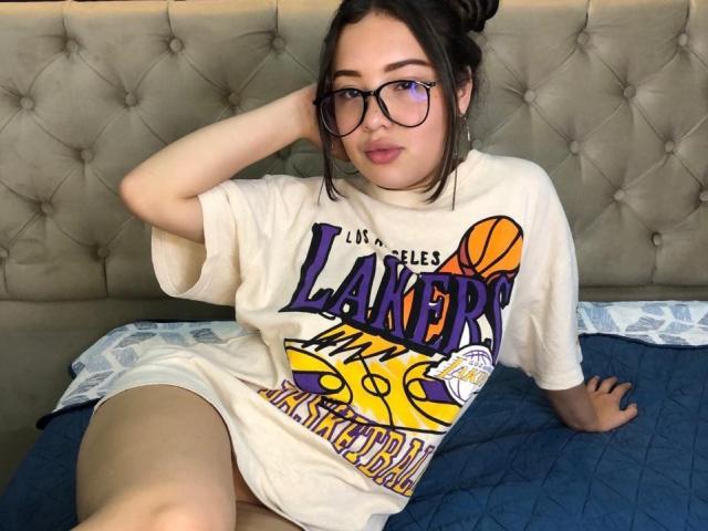 Ryan y Pamela Prince, con gafas, camiseta blanca de Los Angeles Lakers y pantalones cortos blancos, sentadas en una cama azul, con una mano en el pelo, mirando a la cámara, con el trasero ligeramente visible, cabello castaño recogido en un moño..