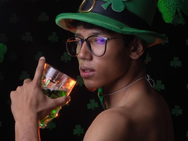 ryanandpamelaprincee1 con gafas, sombrero verde y cuello irlandés, sosteniendo un vaso de whisky, mirando a la cámara, fondo oscuro, cabello negro, boca ligeramente abierta, tréboles verdes..