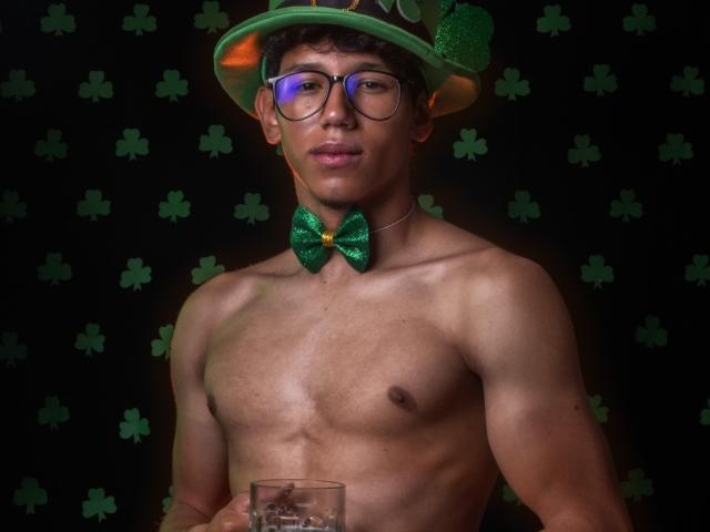 Ryanandpamelaprincee1 ist ein Mann ohne Oberteil, mit einer grünen Fliege und einem Hut, einer Brille, der ein Glas hält und einen muskulösen Brustkorb hat. Er trägt eine grüne, glitzernde Fliege..