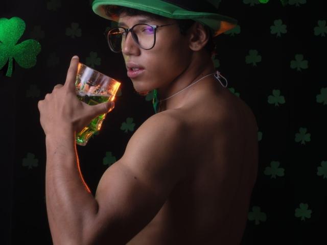 Ryanandpamelaprincee1 with glasses, green hat, and silver necklace, holding a glass of whiskey, showing a muscular, shirtless back, looking over shoulder, dark background with green clovers.