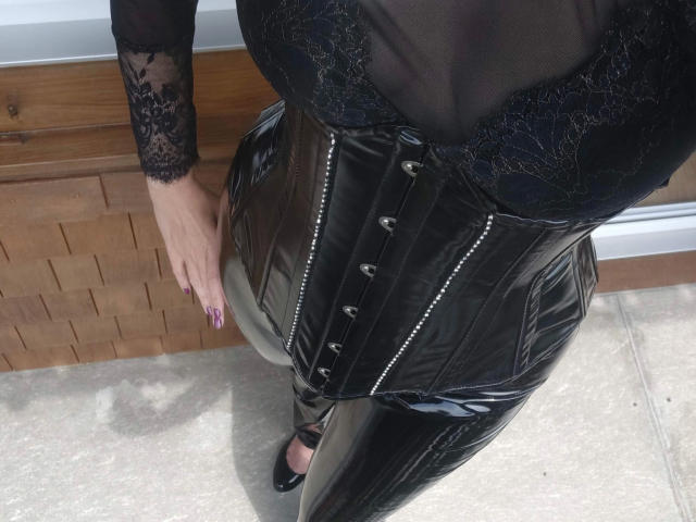 MissShine-hot's black lace top, shiny leather corset, and tight black latex pants, showing her curvy, busty, and butt-filled figure. Her hand with pink nails rests on her side.