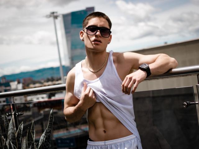 HunterHeatt, muscular with defined abs, wearing a white tank top, lifting it to show his abs, sunglasses, gold necklace, black watch, and white pants, leaning on a railing.