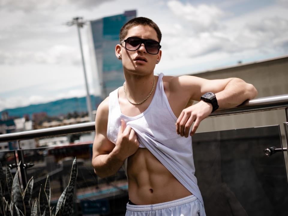 HunterHeatt, muscular with defined abs, wearing a white tank top, lifting it to show his abs, sunglasses, gold necklace, black watch, and white pants, leaning on a railing.