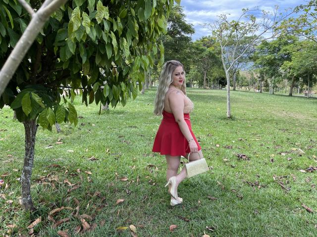 SamanthaGold, avec de longs cheveux blonds, une jupe rouge, un haut en dentelle, des chaussures blanches, tenant un sac beige, debout sur l'herbe, regardant en arrière, un arbre à gauche, un ciel bleu, un parc vert..