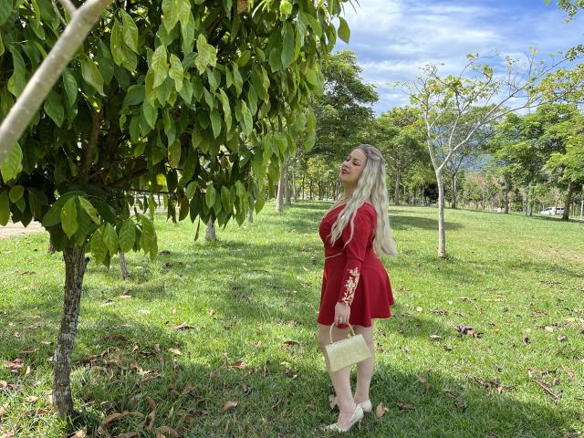 SamanthaGold with long blonde hair, red dress, holding yellow purse, standing under green leafy tree, looking up, grassy park, clear blue sky, wearing white heels.