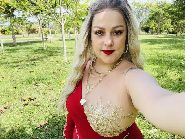 SamanthaGold with blonde hair, red dress, and gold floral embroidery, showing her tits in a selfie, wearing red lipstick and gold jewelry, standing in a grassy park.