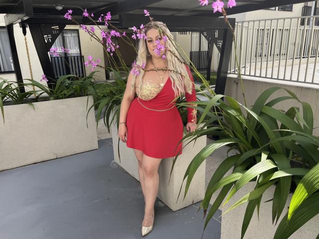 SamanthaGold with long blonde hair, red dress with gold cutout, standing in a patio with purple orchids, wearing beige heels, her tits visible, surrounded by green plants.