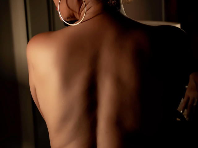 AgathaStone's bare back with visible ribs and soft skin, wearing large hoop earrings, standing in dim light. Her ass is slightly visible from the side.