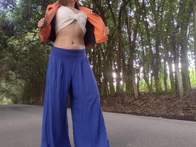 AgathaStone with a flat stomach, orange jacket open, white tied-up shirt, and blue loose pants standing on a road, forest in the background.