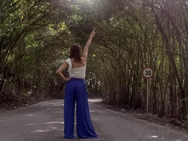 AgathaStone with back to camera, arm raised, wearing white off-shoulder top, blue high-waisted pants, standing on forest road, pointing upward.