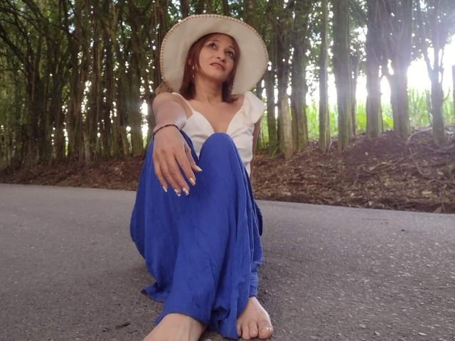 AgathaStone with bare feet, sitting on a road, wearing a white hat, blue dress, and white top, looking up with a slight smile, forest background.