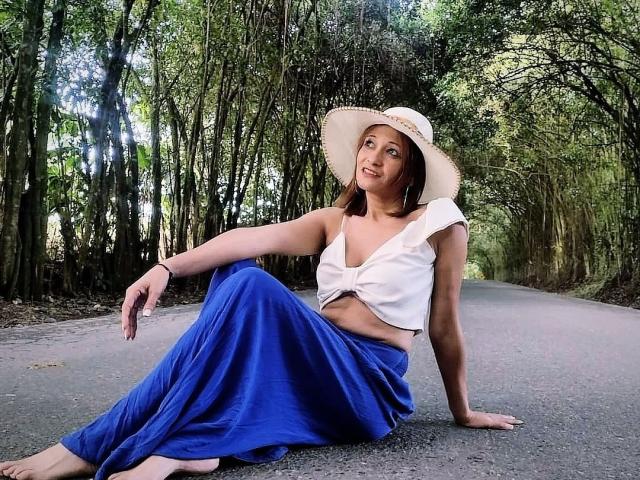AgathaStone, barefoot, wearing a white sunhat, tied-up white top, and blue skirt, sitting on a path with trees, smiling, exposing her midriff and her ass.
