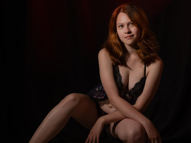 TaylorCarpert with red hair, black lace lingerie, small tits, and a cute smile, sitting with legs crossed, dark background, soft lighting, subtle red glow, sexy, confident.