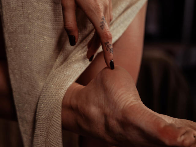 SophieKitty's tattooed finger with black nail polish touches her bare, wrinkled foot. She's lifting a beige, textured dress. Her skin is light, and the focus is on her foot.