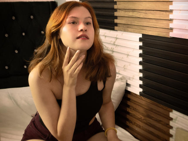 TaylorCarpert has red hair, a black tank top, and is touching her neck with her right hand, wearing maroon shorts, looking slightly confused, with a black cushioned headboard.
