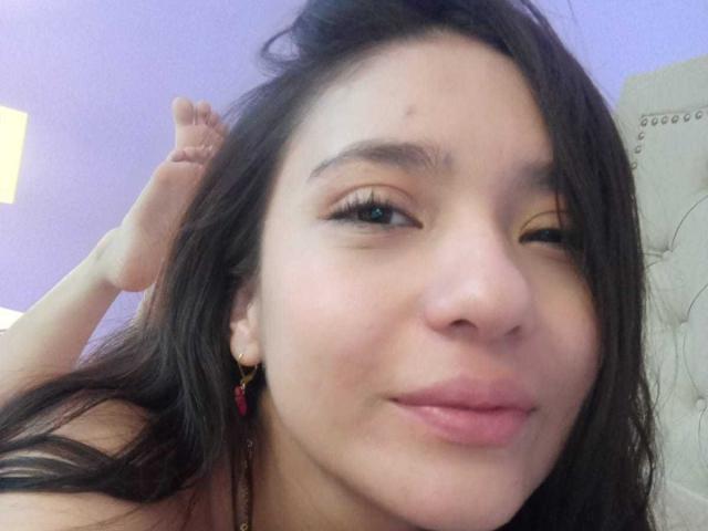 CattsyCathy with brown hair, winking, pink lips, small red earrings, light brown skin, and a slight zit on her cheek, holding her hair up.