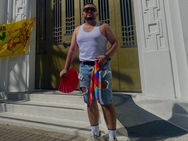 GaelB steht im Vordergrund, trägt ein weißes Tanktop, zerrissene blaue Jeans und Socken, hält einen roten Fächer und ein buntes Band und trägt eine Sonnenbrille..
