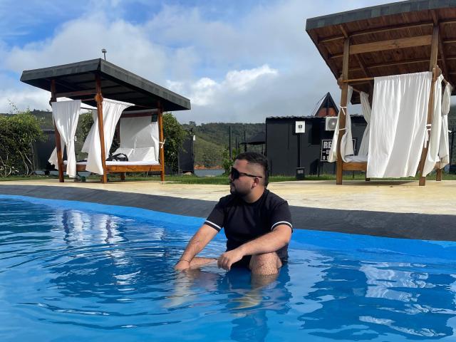 GaelB, avec une barbe et des lunettes de soleil, portant une chemise noire, assis dans une piscine bleue, tenant ses fesses avec de l'eau arrivant jusqu'à ses cuisses, sous un abri en bois au bord de la piscine..