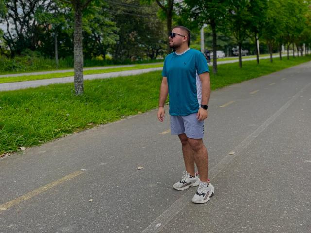GaelB se tient sur un trottoir, portant une chemise turquoise, un short gris, des baskets blanches, des lunettes de soleil noires et une montre noire. Il regarde sur le côté avec une posture détendue..