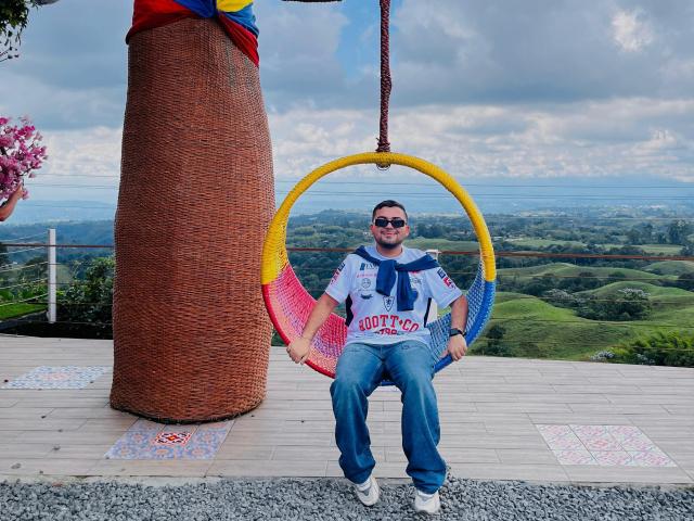 GaelB sentado en un columpio circular amarillo y rojo, vistiendo una camiseta con la inscripción "Boo-Tay", jeans azules y gafas de sol, con un gran pilar de mimbre a la izquierda y una vista panorámica..