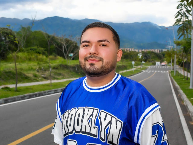 GaelB, avec des cheveux bruns courts et une barbe, portant un maillot de football bleu et blanc, debout sur une route, souriant, avec des montagnes en arrière-plan, et un ciel légèrement nuageux..