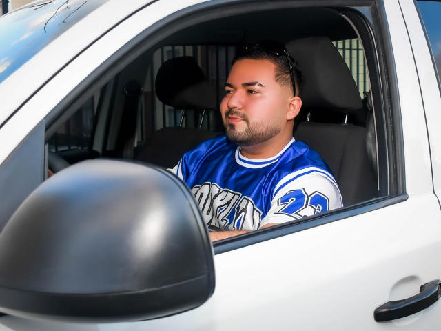 GaelB, ein etwas korpulenter Mann mit kurzem Bart, sitzt in einem weißen Auto, trägt ein blau-weißes Fußballtrikot und blickt mit ernstem Gesicht aus dem Fenster..