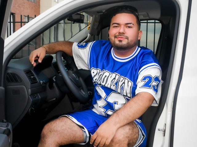 GaelB, un homme corpulent avec une barbe, est assis dans une voiture blanche, portant un maillot de basket bleu et blanc, avec le numéro 24 et le mot "Pacific" dessus, et il a l'air détendu..
