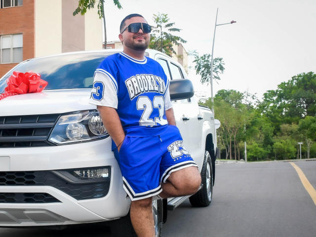 GaelB, un hombre con barba y piel oscura, que lleva una camiseta y pantalones cortos azules y blancos de Bruisers 23, apoyado contra una camioneta blanca, sonriendo, con gafas de sol, y con las manos en los bolsillos..