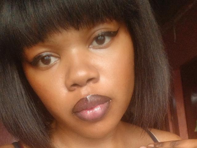 KatyBTeresa has dark brown skin, straight black hair with bangs, and shiny purple lips. She's looking slightly to the side with a small drop on her lower lip.