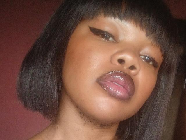 KatyBTeresa has shiny brown skin, black straight bob, and is sticking out her glossy purple-tinted tongue with black winged eyeliner. Her lips are slightly parted.